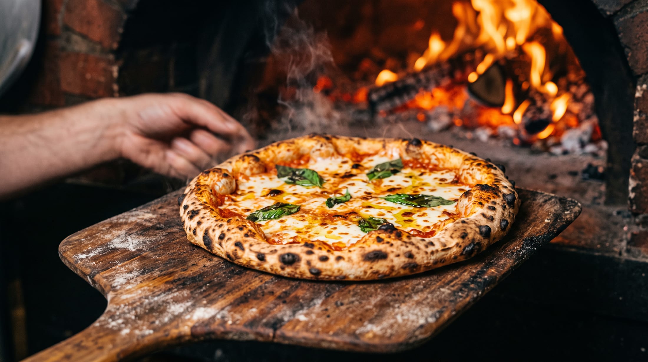 Pizza napoletană scoasă pe lopată din cuptorul pe lemne, cu flăcări în spate